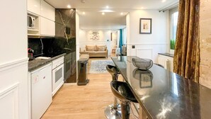 A modern kitchen with a central island, a dining area with a glass table, and a living room with a sofa and a painting on the wall. - UrbanFlat 113 - Groundfloor Apartment Paris Historic Center (París)