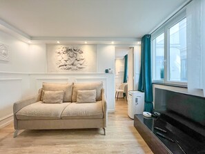 A living room with a beige sofa, a wall-mounted artwork, a flat-screen TV, and a window with curtains. - UrbanFlat 113 - Groundfloor Apartment Paris Historic Center (París)