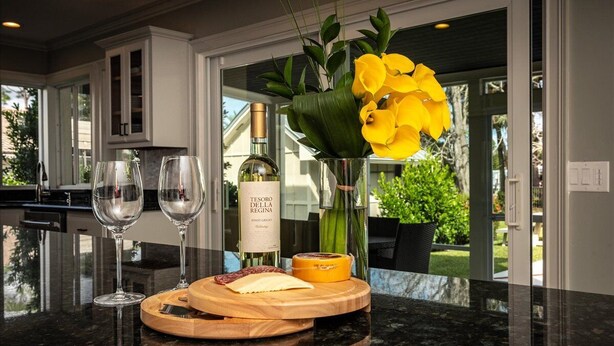 A kitchen counter with two wine glasses, a bottle of wine, a cheese board with cheese and a jar of jam, and a vase with yellow flowers. - Paradise Palm River House w/  WATERFRONT OCEAN ACCESS & PRIVATE POOL AND DOCK (Stuart)