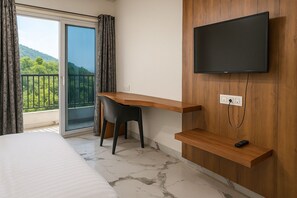 A hotel room with a bed, a desk with a chair, a TV mounted on a wooden wall, and a balcony with a view of greenery. - HOTEL KARDHAR INN (Ambaji)