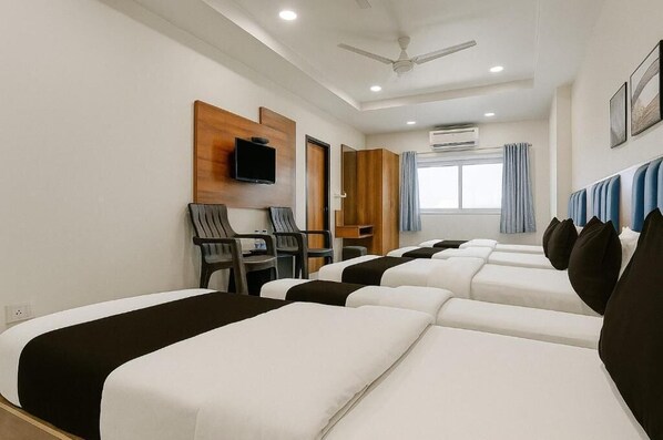 A hotel room with four beds arranged in a row, a desk with chairs, a television mounted on the wall, and an air conditioning unit. - HOTEL KARDHAR INN (Ambaji)