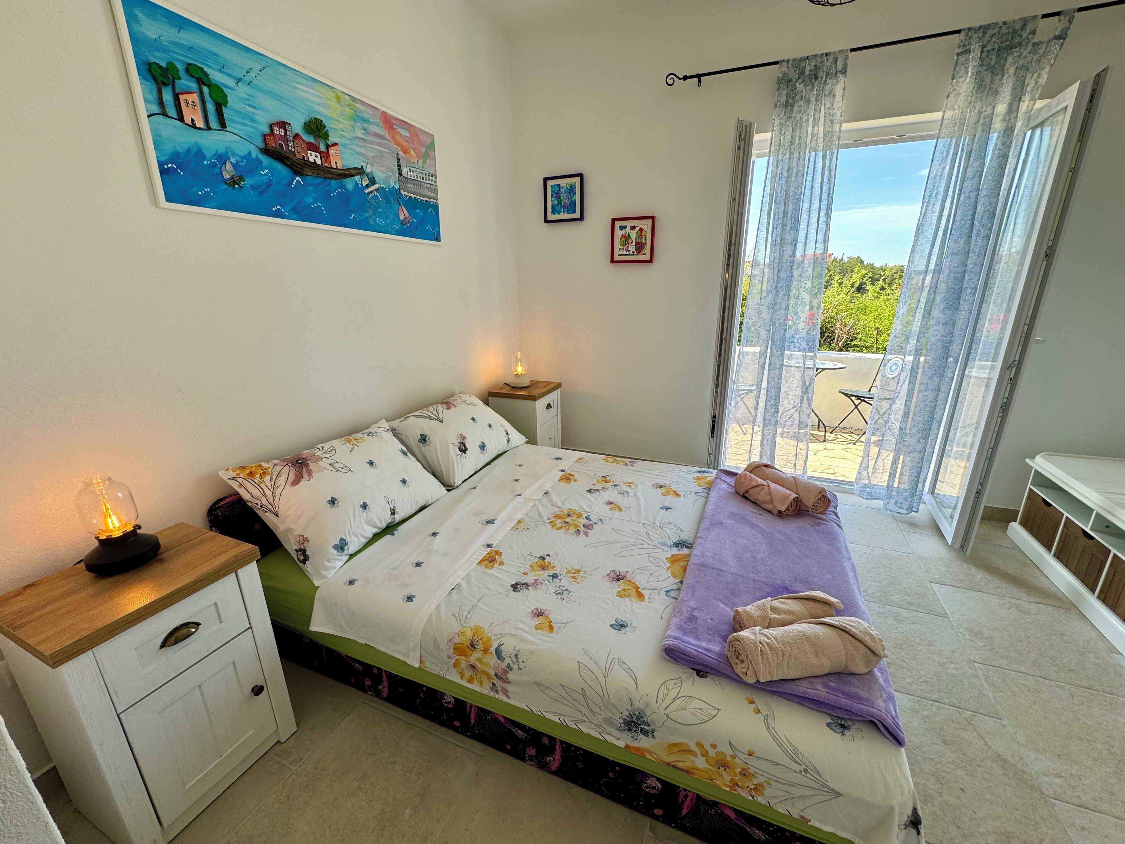 A bedroom with a bed, a nightstand, a bedside lamp, a window with curtains, and a painting on the wall.