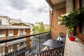 Een balkon met een tafel en stoelen, een bloempot en uitzicht op omliggende gebouwen.