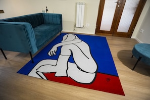 A room with a teal sofa, a blue and red rug with a white abstract design, a radiator, and a wooden door. - Scena Illustrata-Redi 51-un to 10 people (Florence)