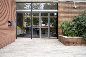 A building entrance with glass doors, a mailbox, and a brick planter with plants. - Scena Illustrata-Redi 51-un to 10 people (Florence)