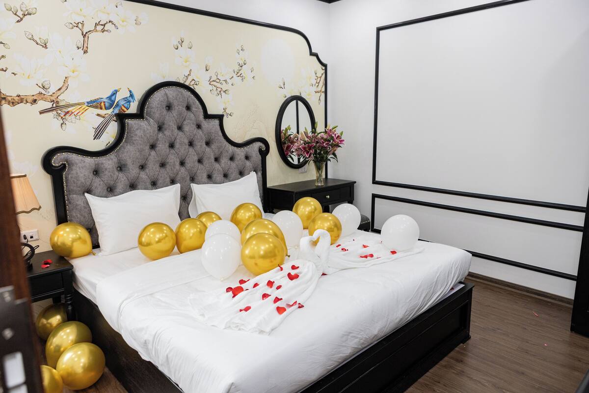 Un lit bien fait, décoré de ballons blancs et dorés, un miroir et un mur orné de motifs floraux et d’oiseaux.
