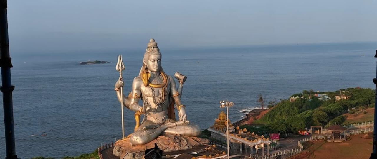 A large stone statue of a deity with multiple arms, seated on a rock with a view of the sea and a small shrine in the background.