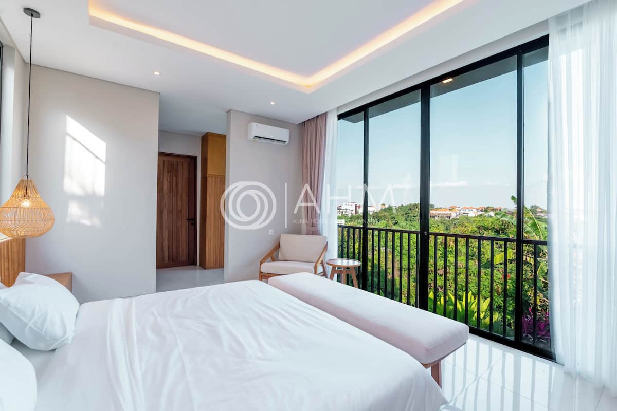 A modern hotel room with a large bed, a small sofa, a balcony with a view of greenery, and a hanging light fixture.