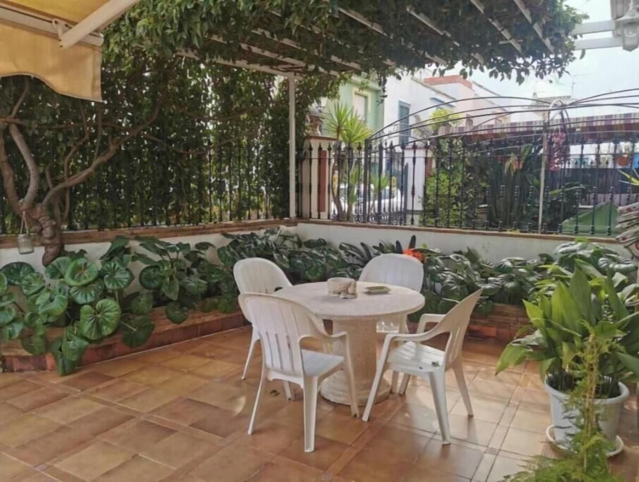 Um pátio com uma mesa redonda e quatro cadeiras, cercado por plantas verdes exuberantes e um piso de azulejos.