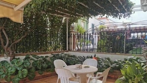 A patio with a round table and four chairs, surrounded by lush green plants and a tiled floor. - Semi-detached house - Los Boliches - Fuengirola (FUENGIROLA)