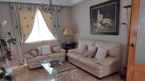 A living room with two beige sofas, a glass coffee table, a chandelier, a painting on the wall, and a window with curtains. - Semi-detached house - Los Boliches - Fuengirola (FUENGIROLA)