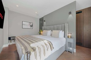 Ein modernes Schlafzimmer mit einem großen Bett, einem Nachttisch mit Lampe, einem an der Wand montierten Fernseher und einem gerahmten Bild.