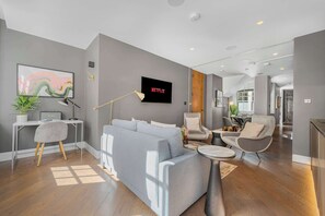 Ein modernes Wohnzimmer mit einem grauen Sofa, einem Schreibtisch mit Lampe, einem Fernseher, der 'NETFLIX' anzeigt, und einem Essbereich mit Stühlen.