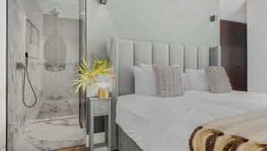 A modern bedroom with a large bed, a nightstand, a glass shower enclosure, and a potted plant.