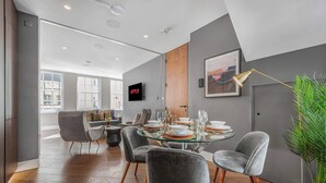 A modern dining area with a glass table set for six, grey upholstered chairs, a mounted television displaying 'NETFLIX', and a large window providing natural light.