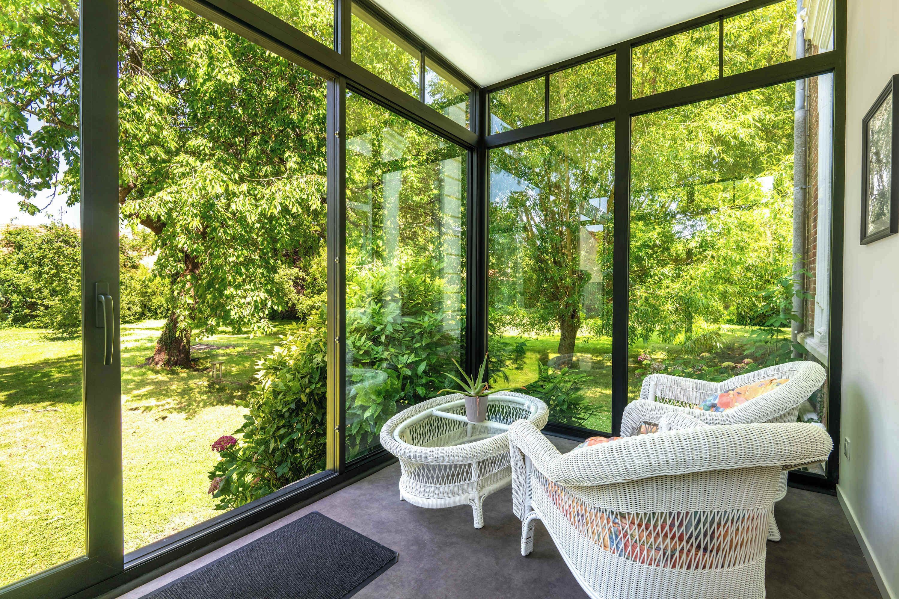 A conservatory with large glass windows, wicker furniture, and a view of a lush garden.