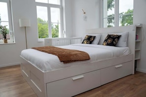 Un lit blanc avec une couverture marron, des oreillers blancs et une table de chevet avec une lampe.