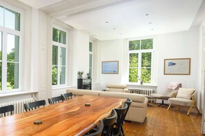 A spacious room with a large wooden dining table, multiple chairs, a sofa, and two windows with views of greenery.