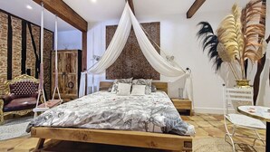 Un dormitorio con una cama de madera, una silla colgante, una mesita de noche, un tapiz decorativo en la pared y un florero con plumas.