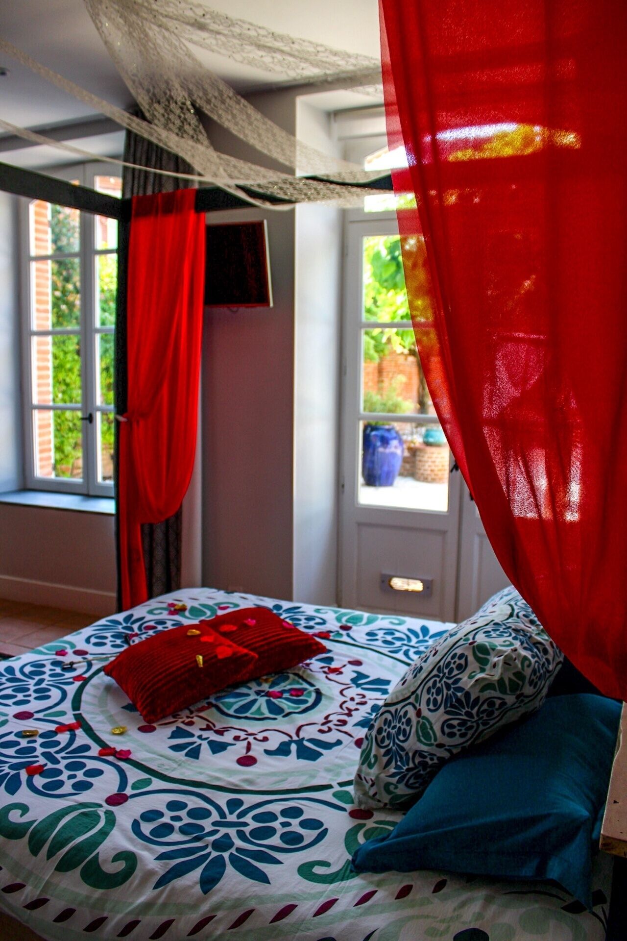A bedroom with a bed, red curtains, a patterned bedspread, and a window with a view of greenery.