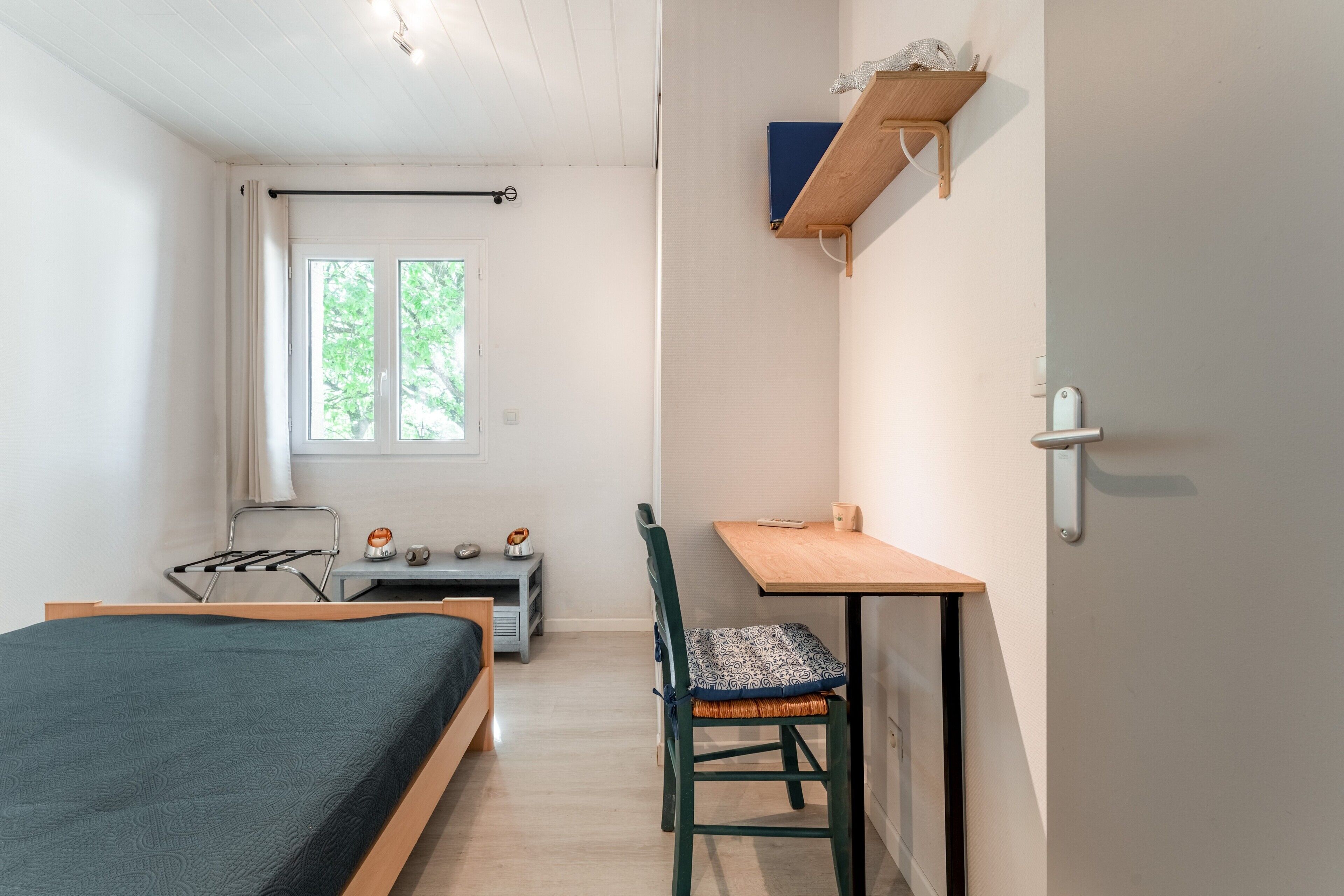 A small, well-lit room with a bed, a desk, a chair, a window, and a shelf with a book.