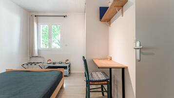 A small, well-lit room with a bed, a desk, a chair, a window, and a shelf with a book.