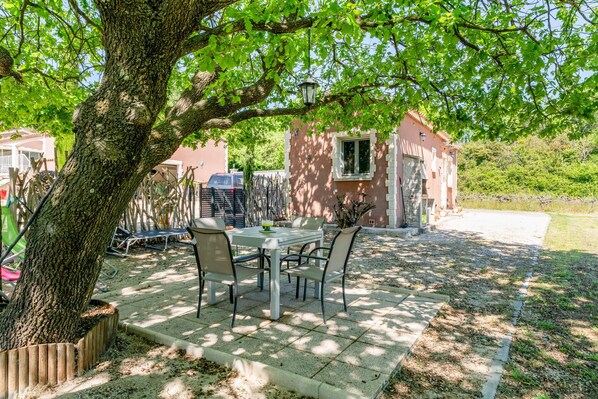 A patio with a table and chairs, a tree, and a small building in the background. - La Petite Maison in the Vineyard (Gaujac)