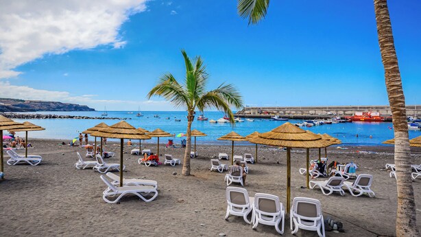 Una playa con sillones blancos y sombrillas de paja, un cielo azul claro y barcos en el agua.