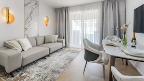 En moderne stue med sofa, spisebord, stole og et stort vindue med gardiner.