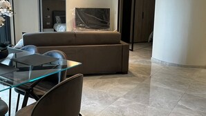 A modern living room with a glass dining table, dark chairs, a beige sofa, and a large TV.