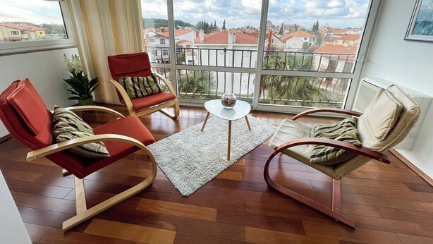 Un salon moderne doté d’un grand balcon, d’un parquet en bois et de fauteuils rouges et beiges.
