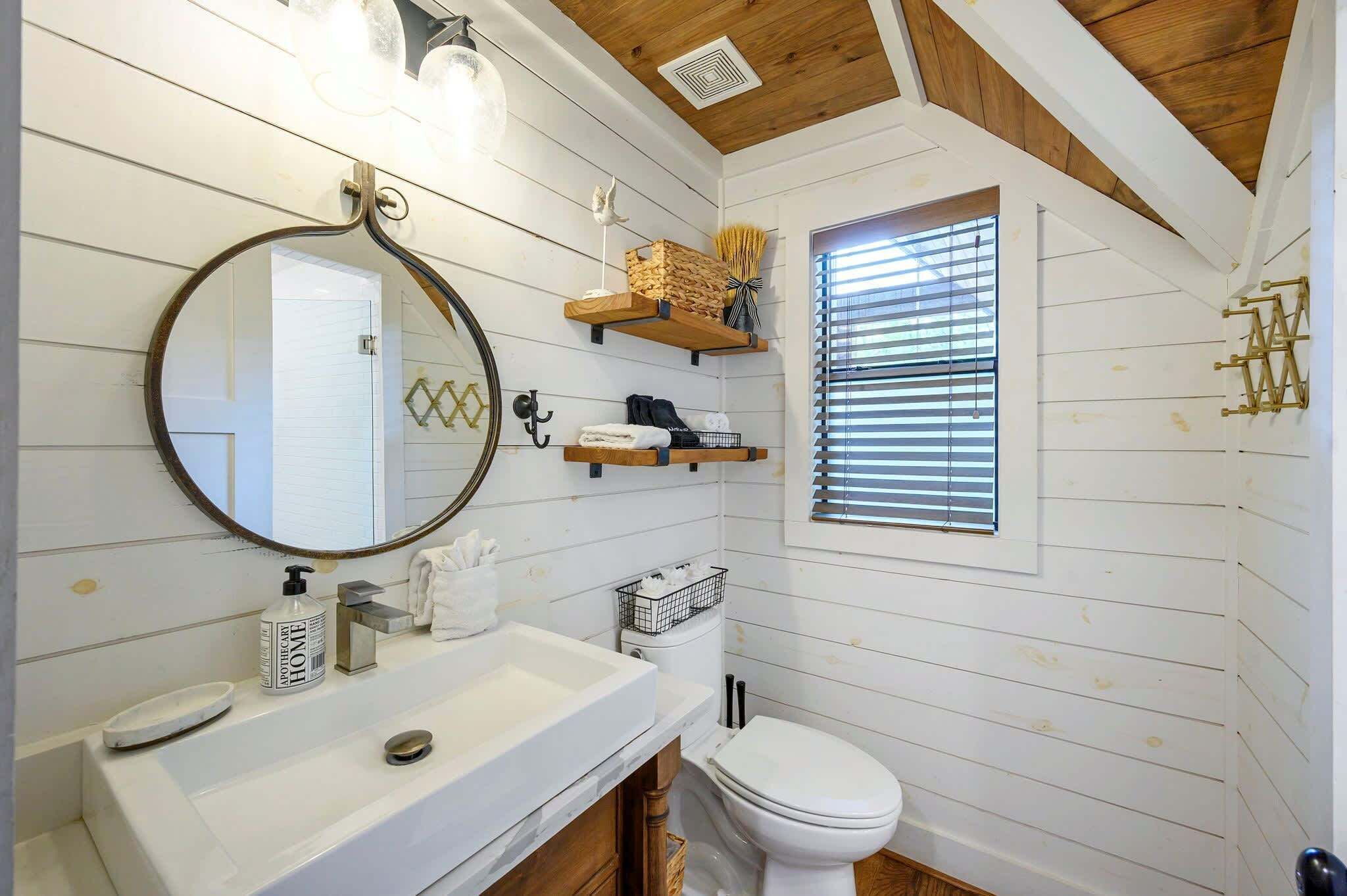 A bathroom with a wooden ceiling, a round mirror, a white sink, a toilet, and a window with blinds.