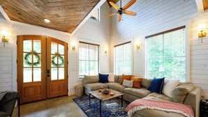 A spacious living room with a wooden ceiling, a ceiling fan, a large window with blinds, a beige sectional sofa, and a wooden coffee table. - Sugarberry Farm Farmhouse! Firepit, Porch Swing (Broken Bow)