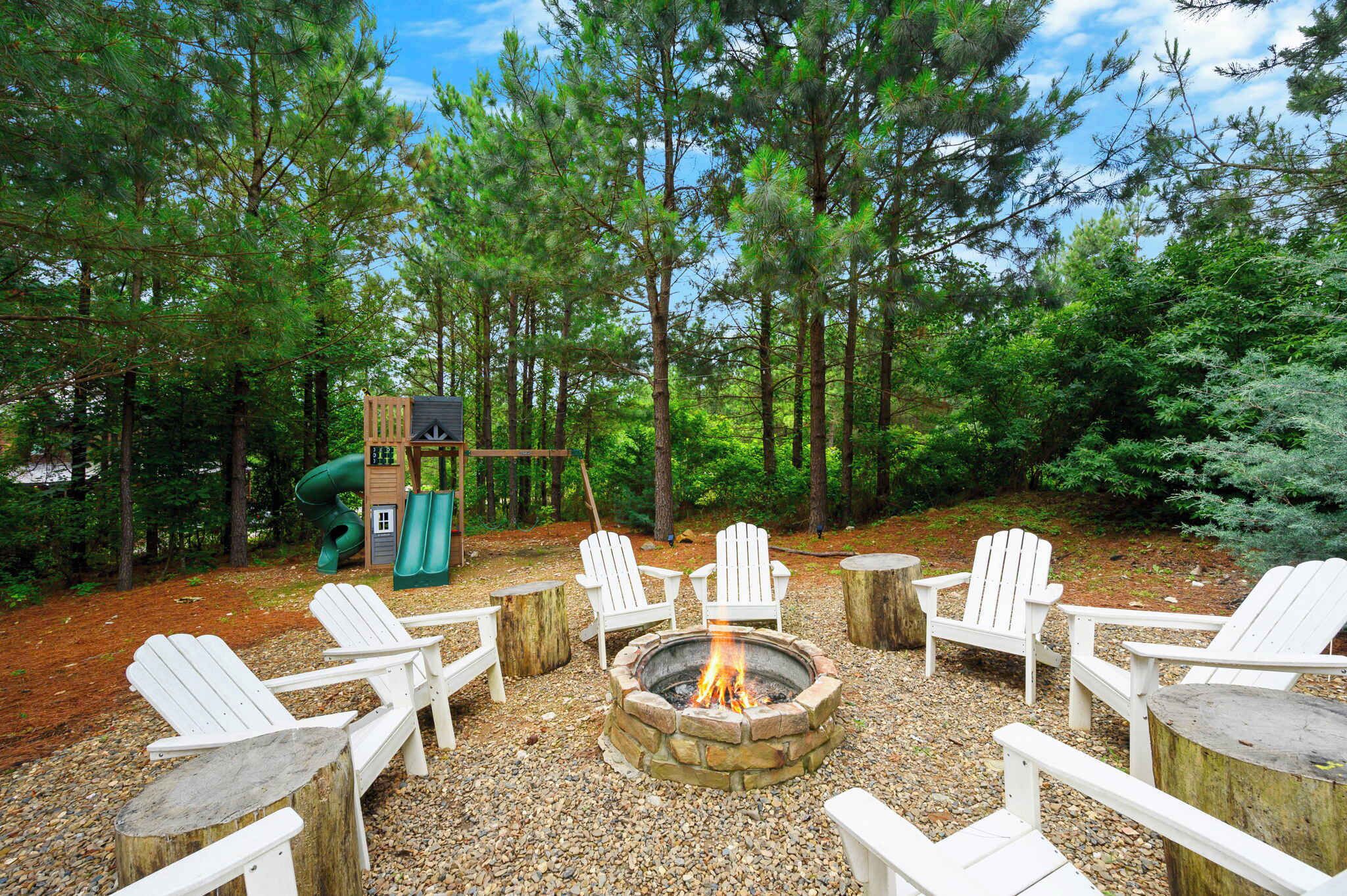 A fire pit surrounded by Adirondack chairs, a play structure, and a forested area.