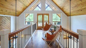 A wooden interior with a high ceiling, large windows, and a balcony with chairs and a table. - Sugarberry Farm Farmhouse! Firepit, Porch Swing (Broken Bow)