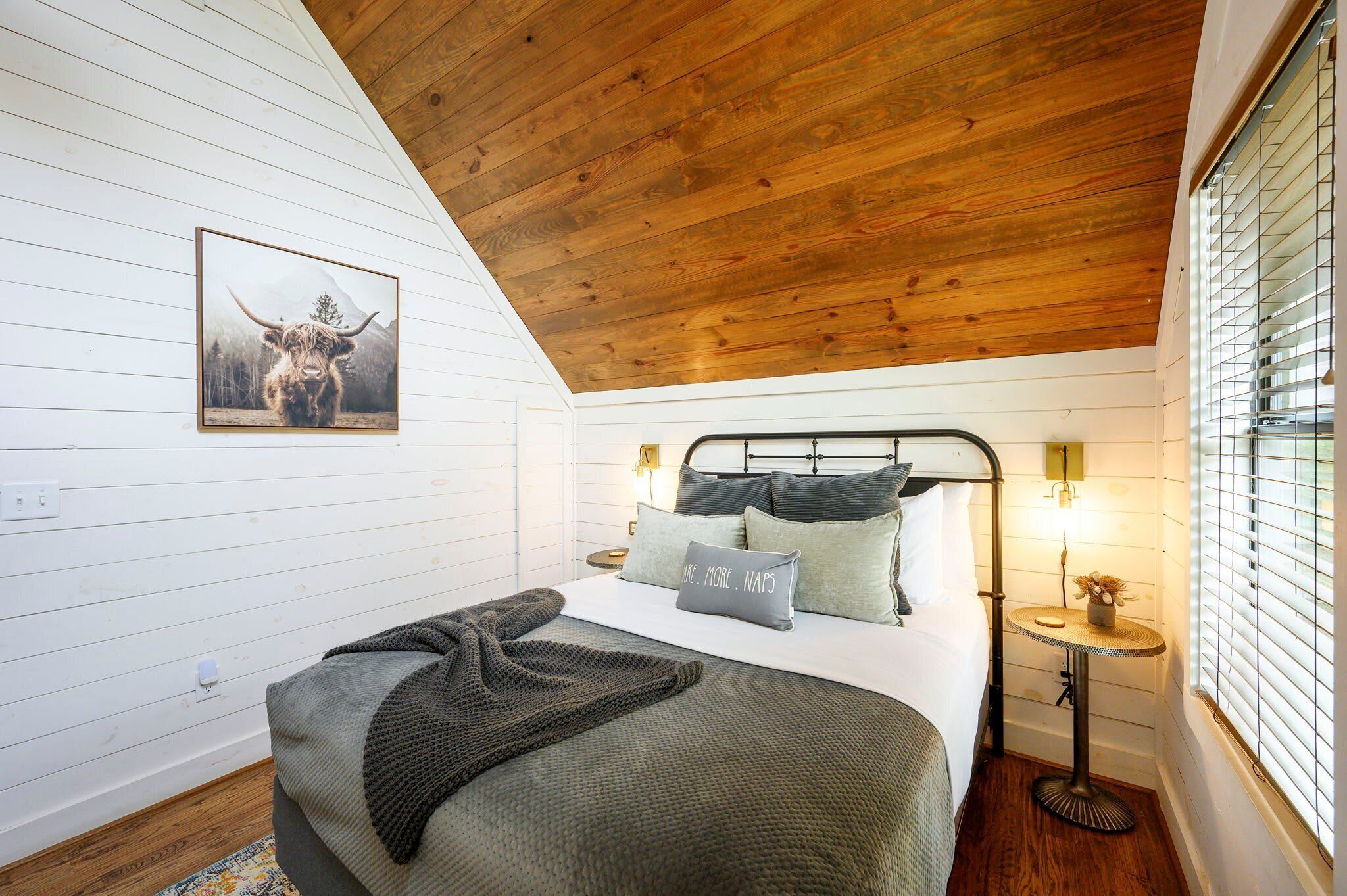 Een slaapkamer met een schuine plafond, een bed met kussens, een nachtkastje met een lamp en een lijst met een afbeelding aan de muur.