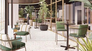 Un espace de détente intérieur moderne, avec des chaises et des tables à revêtement vert, entouré de grands pots de plantes et séparé par une cloison en verre.