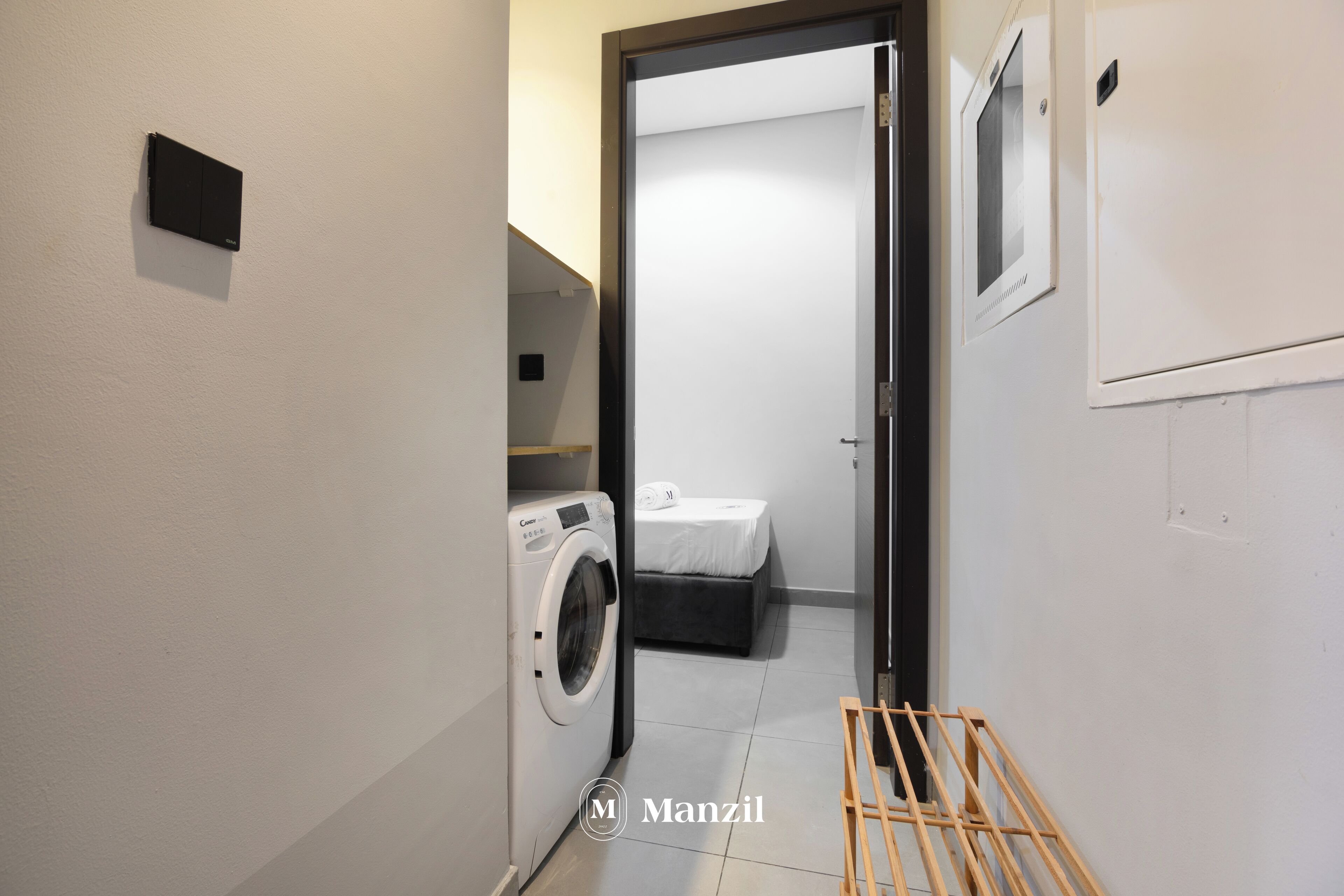 A laundry room with a washing machine, a bed, and a small wooden drying rack.
