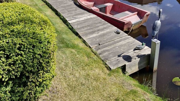 A calm canal with a wooden dock, a red boat, and a small building. - Haus 53° Nord - North Sea vacation for the whole family - pets welcome (Südbrookmerland)