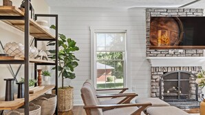 A cozy living room with a stone fireplace, wooden furniture, and a large window overlooking a garden. - Call Me Old Fashioned! *Heart of Bardstown Square* (Bardstown)