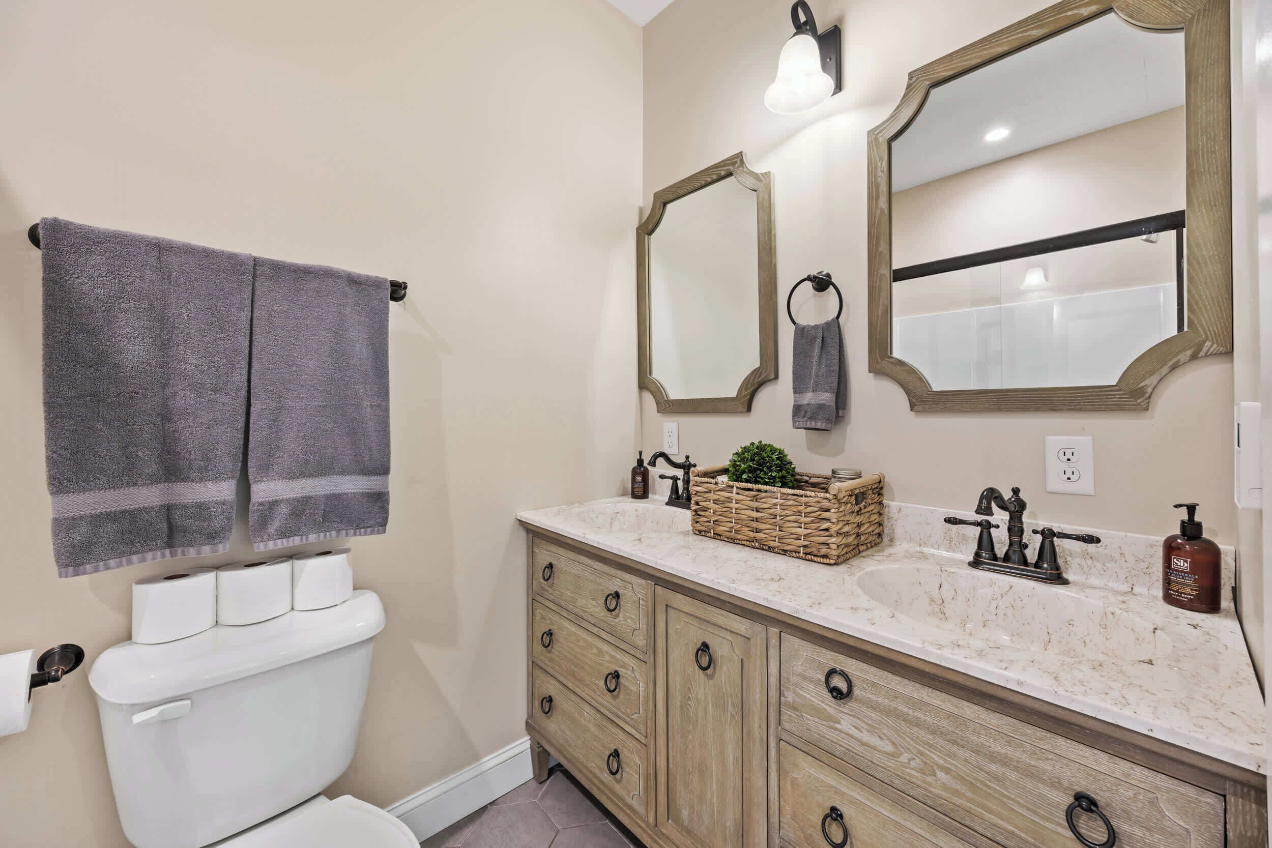 A bathroom with a toilet, two mirrors, a sink with a marble countertop, and wooden cabinets.