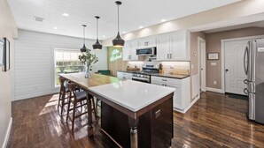 A modern kitchen with white cabinets, a central island with bar stools, a stainless steel refrigerator, and a large window providing natural light. - Call Me Old Fashioned! *Heart of Bardstown Square* (Bardstown)