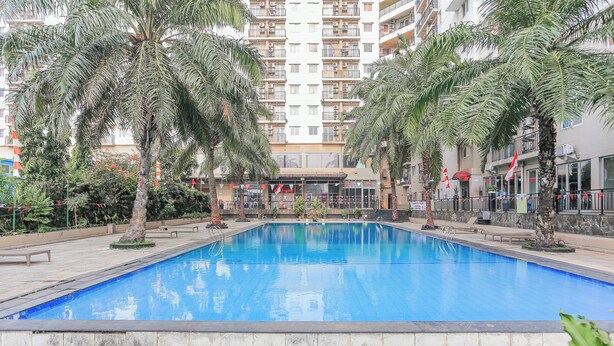 A swimming pool surrounded by palm trees and a multi-story building in the background. - Capital O 94796 Apartment Mutiara Bekasi (Bekasi)