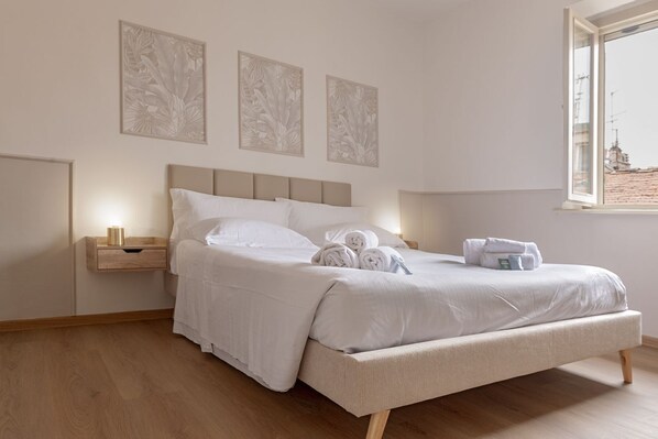 A neatly made bed with white linens and rolled towels, a bedside table with a lamp, and three wall-mounted artworks.
