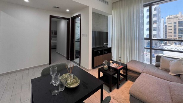 A modern living room with a sofa, a dining table with glasses and a plate, a TV, and a window with a city view. - Luxury 1-bedroom fully furnished Lifeline apartment  (Abu Dhabi)