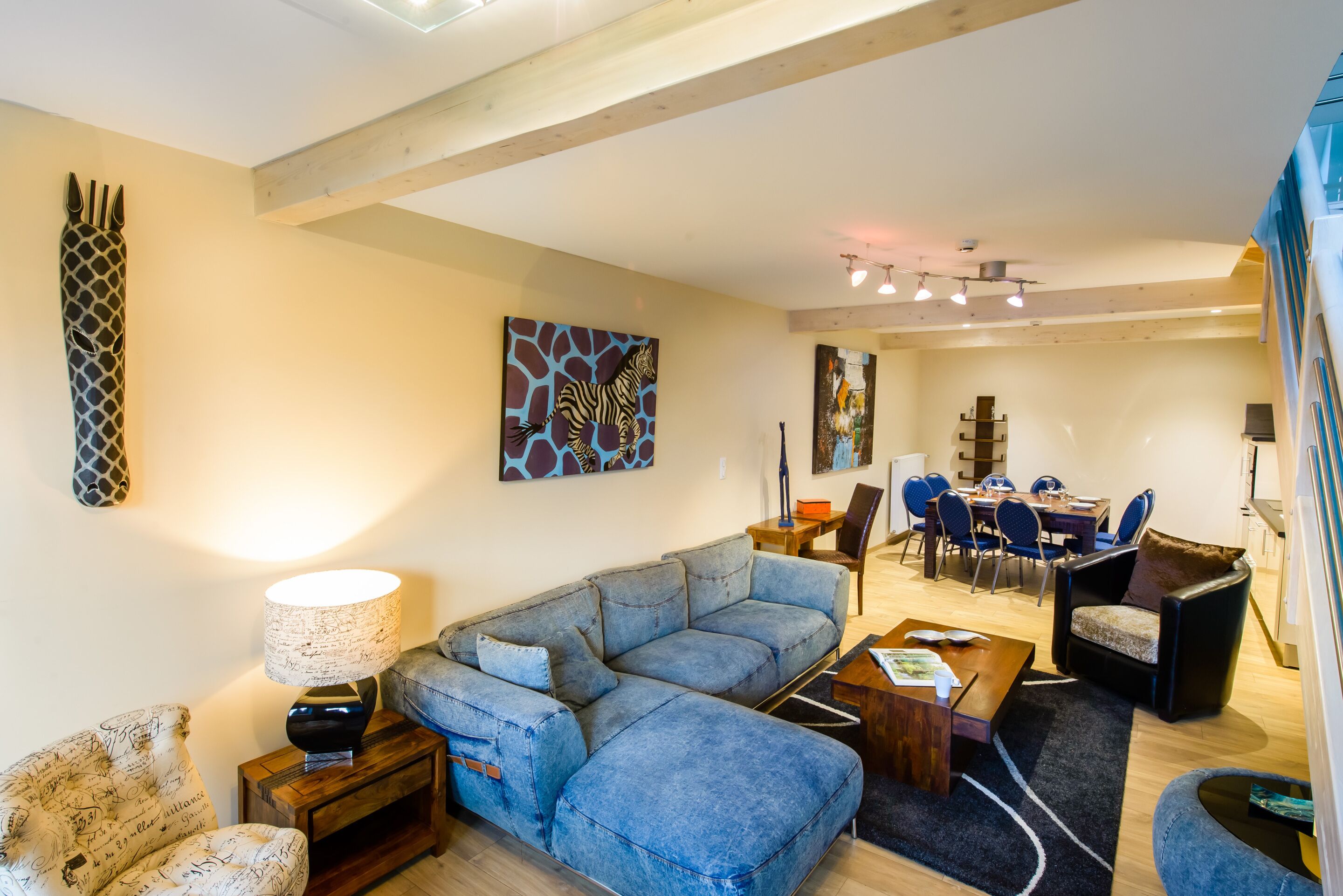 A modern living room with a sectional sofa, a dining area with blue chairs, and a kitchen in the background.