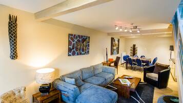 A modern living room with a sectional sofa, a dining area with blue chairs, and a kitchen in the background.