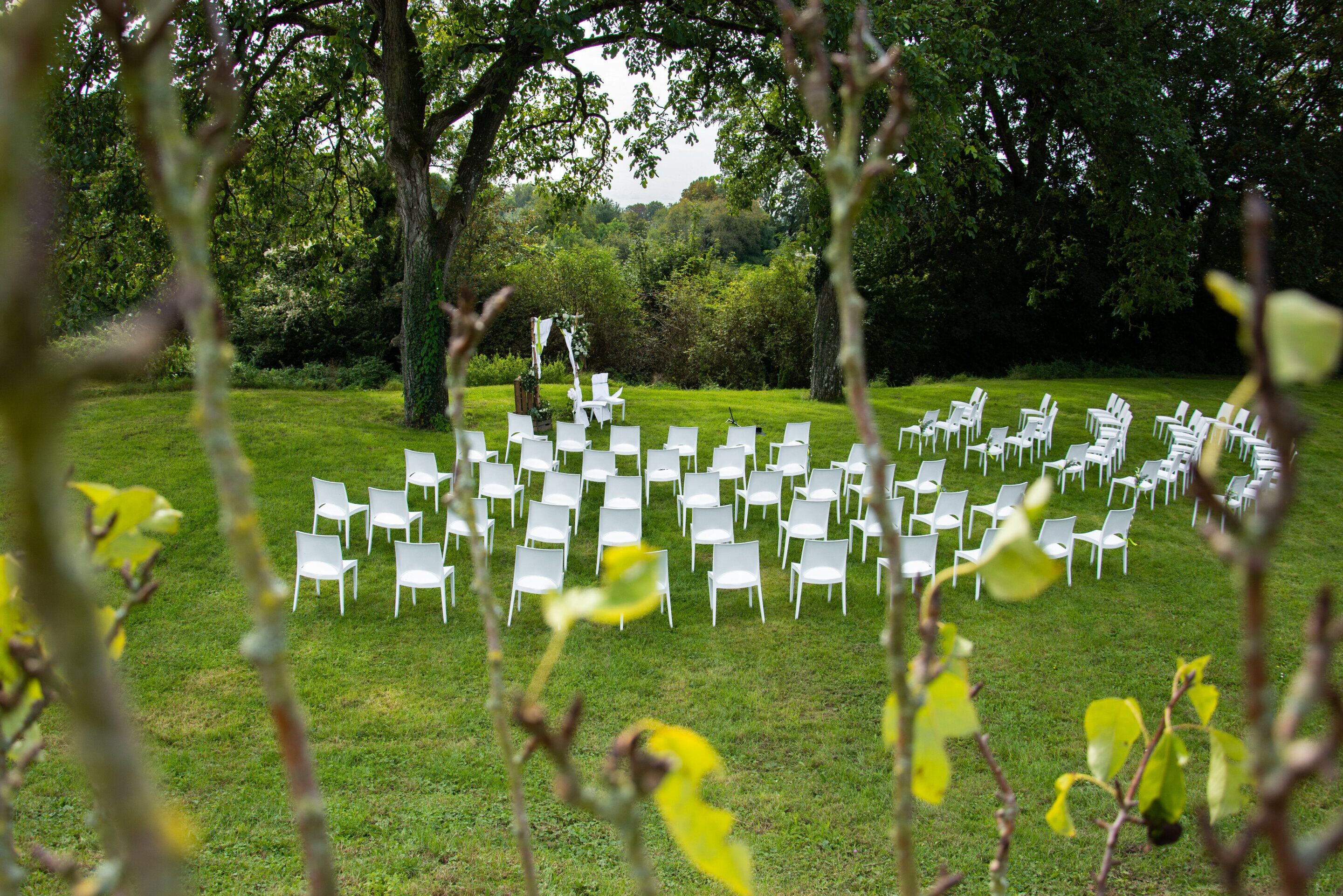 An outdoor seating area with white chairs arranged in rows, surrounded by trees and greenery.