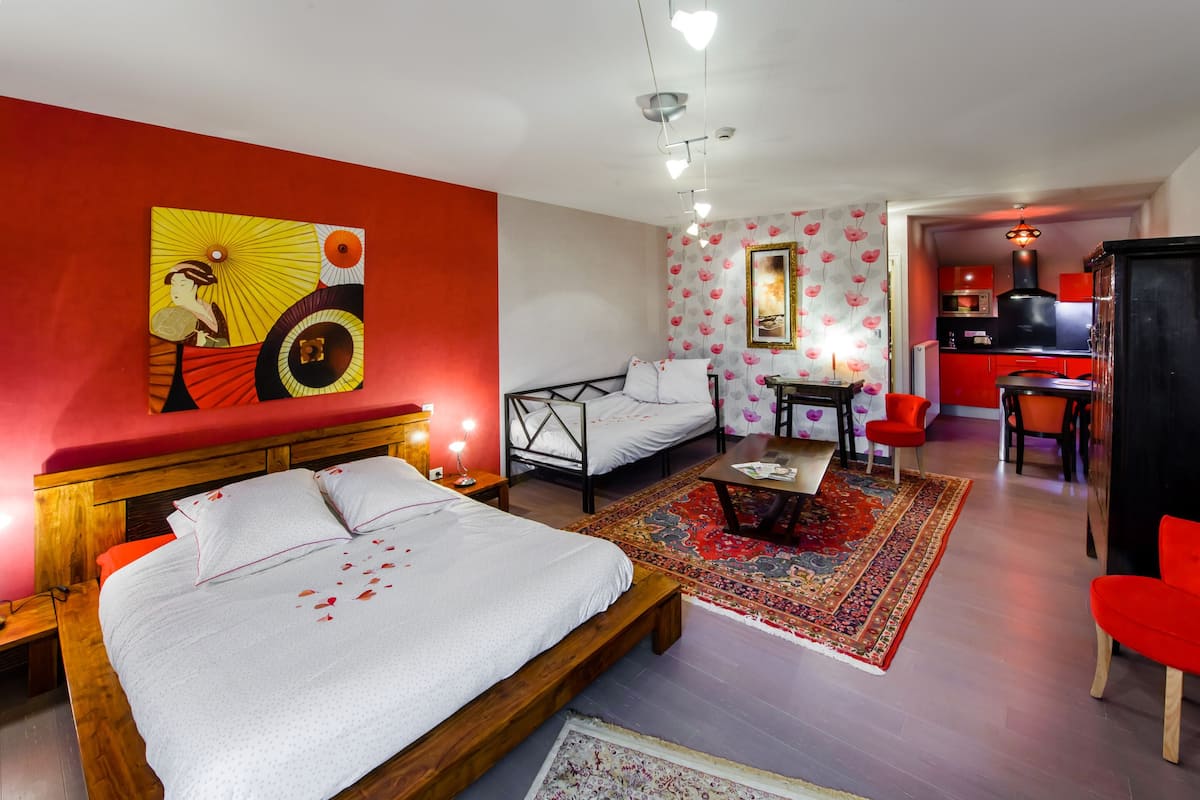 Une chambre à coucher comprenant un lit, des tables de chevet, une commode, un bureau, une chaise, un canapé rouge, un fauteuil rouge, un tapis rouge, un mur rouge, un luminaire rouge au plafond, un miroir avec un cadre rouge, un tableau avec un cadre rouge et un coin cuisine avec un cadre rouge.