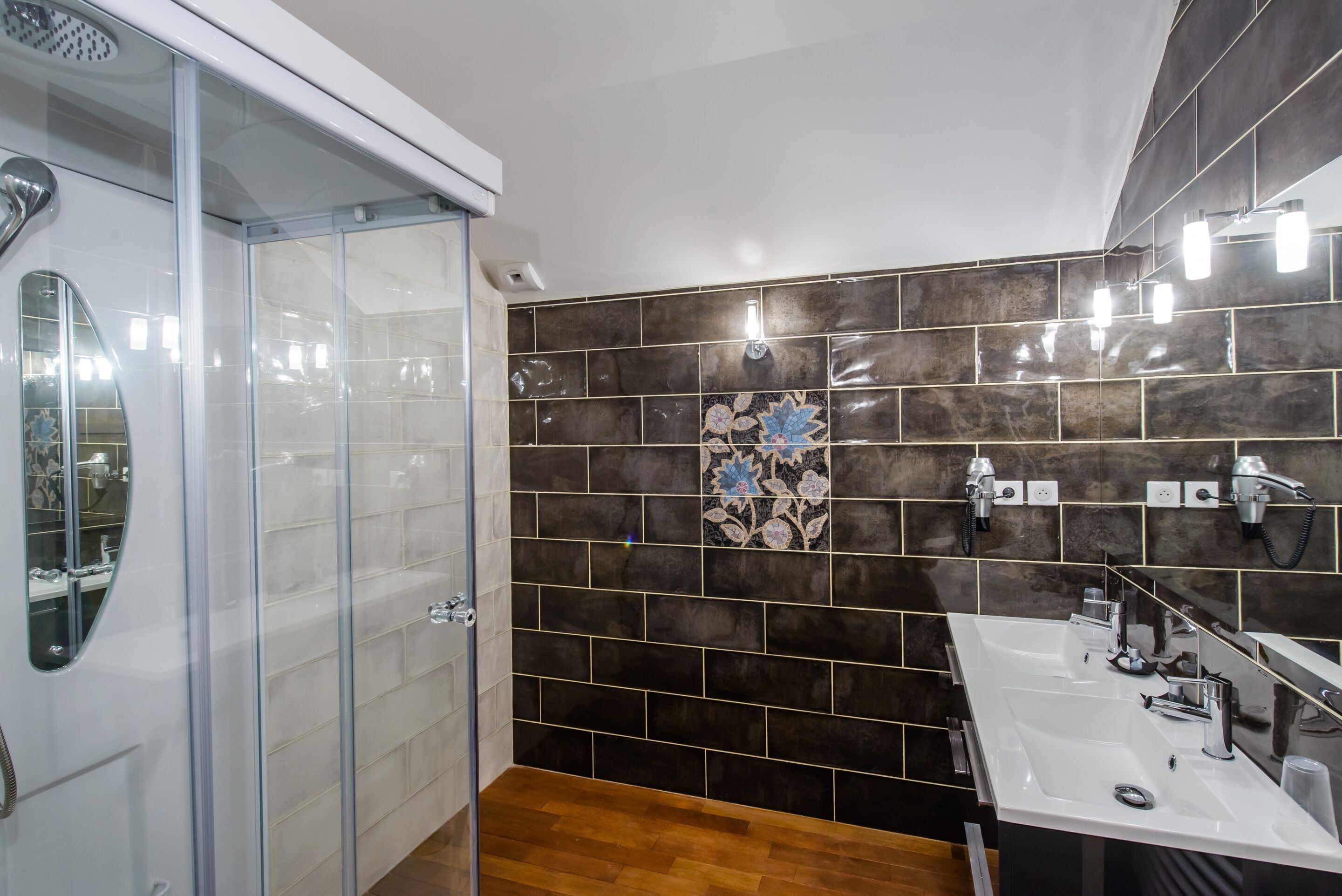 Un baño moderno con cabina de ducha de vidrio, un lavamanos espacioso con espejo y paredes de azulejos oscuros.